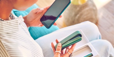 A woman holding a smart phone in her left hand and an IQOS device in her right hand.