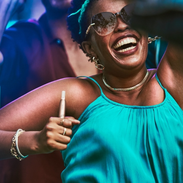 Woman dancing and smiling with a stellar blue IQOS 3 DUO holder in her hand.