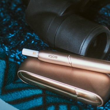 Gold IQOS holder and charger on blue patterned fabric next to binoculars