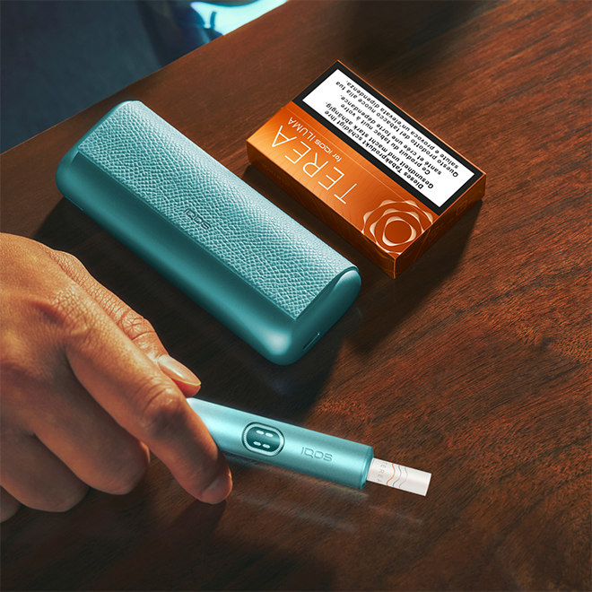 Hand holding IQOS ILUMA i PRIME device, with charger and pack of TEREA tobacco sticks on a table