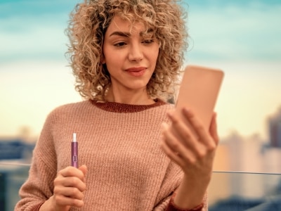 Frau schaut auf ihr Smartphone, während sie ein IQOS ILUMA i PRIME Gerät hält