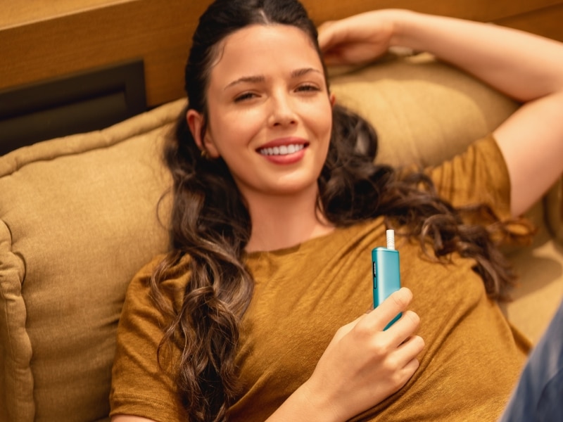 Frau liegt auf der Couch und hält ein IQOS ILUMA i ONE Gerät
