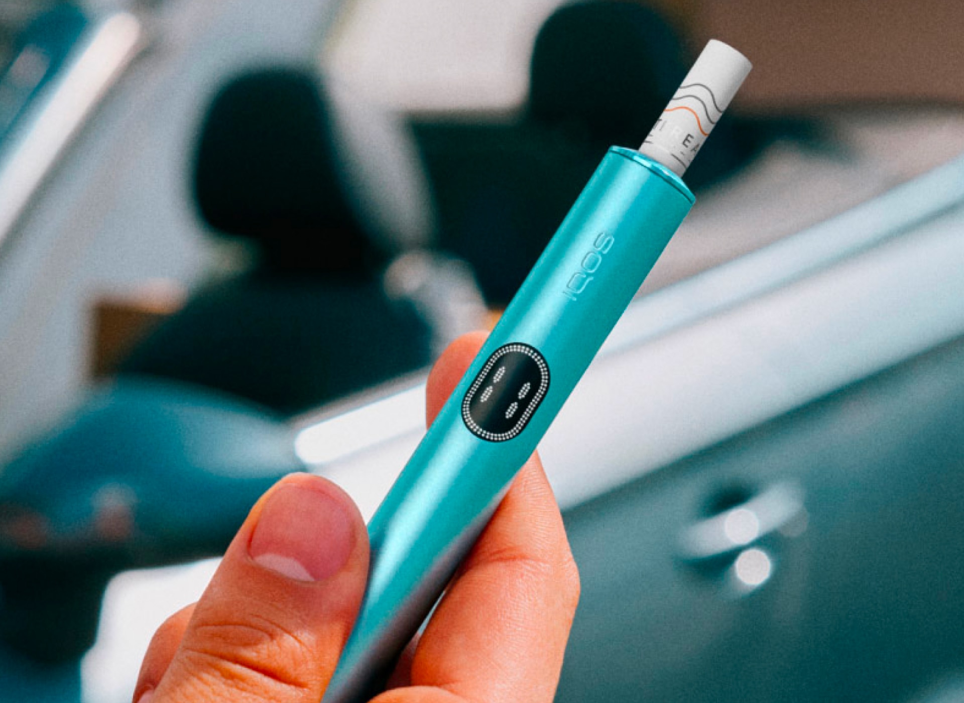 Main tenant un appareil IQOS ILUMA i bleu avec une voiture en arrière-plan.
