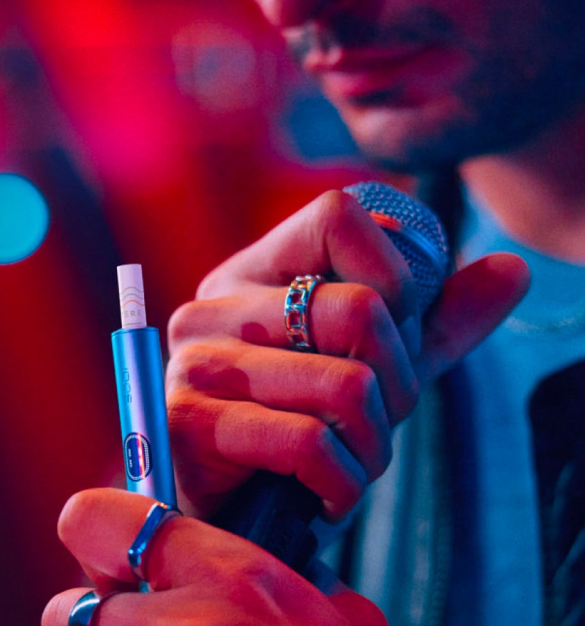 Un homme tenant un appareil IQOS ILUMA i bleu et un micro avec un rideau rouge en arrière-plan.