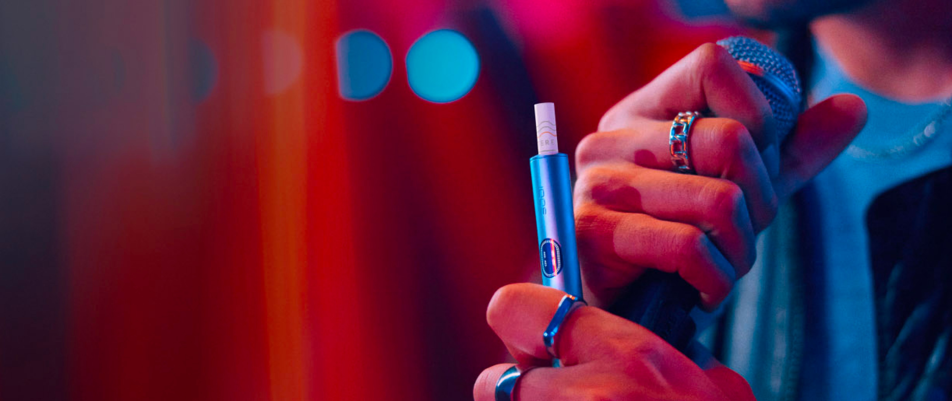 Mann mit einem blauen IQOS ILUMA Gerät und Mikrofon, im Hintergrund ein roter Vorhang.